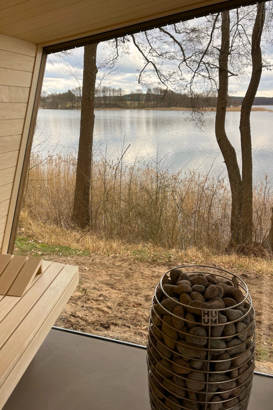 Der Blick aus einer modernen Sauna mit Steinofen durch ein großes Fenster auf einen See und Bäume – der perfekte Rückzugsort nach einem Tag Winter- oder Herbstcamping, an dem die ruhige Landschaft Ihr saisonales Abenteuer noch verstärkt.