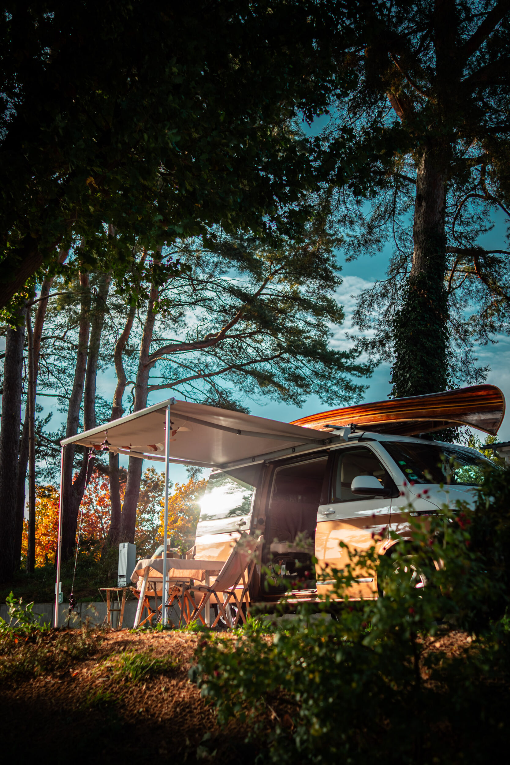 Ein Wohnmobil mit Markise und Stühlen steht auf einer sonnendurchfluteten Waldlichtung und bildet den perfekten Ausgangspunkt für entspannte Herbstcamping-Abenteuer.