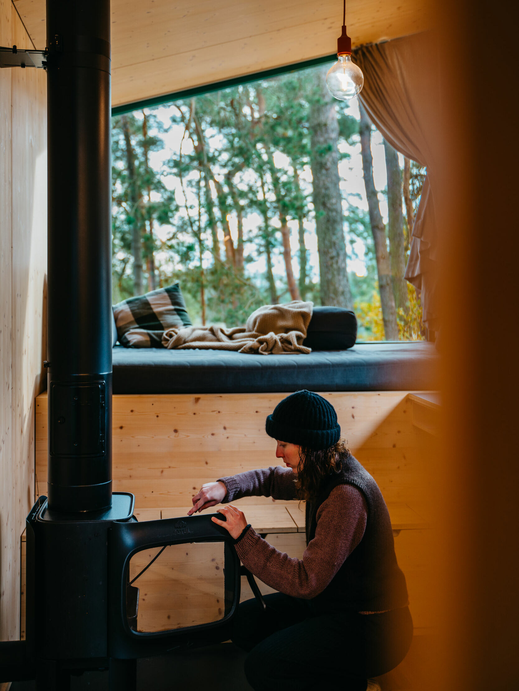Eine Person mit einer Mütze heizt einen Holzofen in einer gemütlichen Hütte mit einer Fensterbank und Blick auf den Wald an.