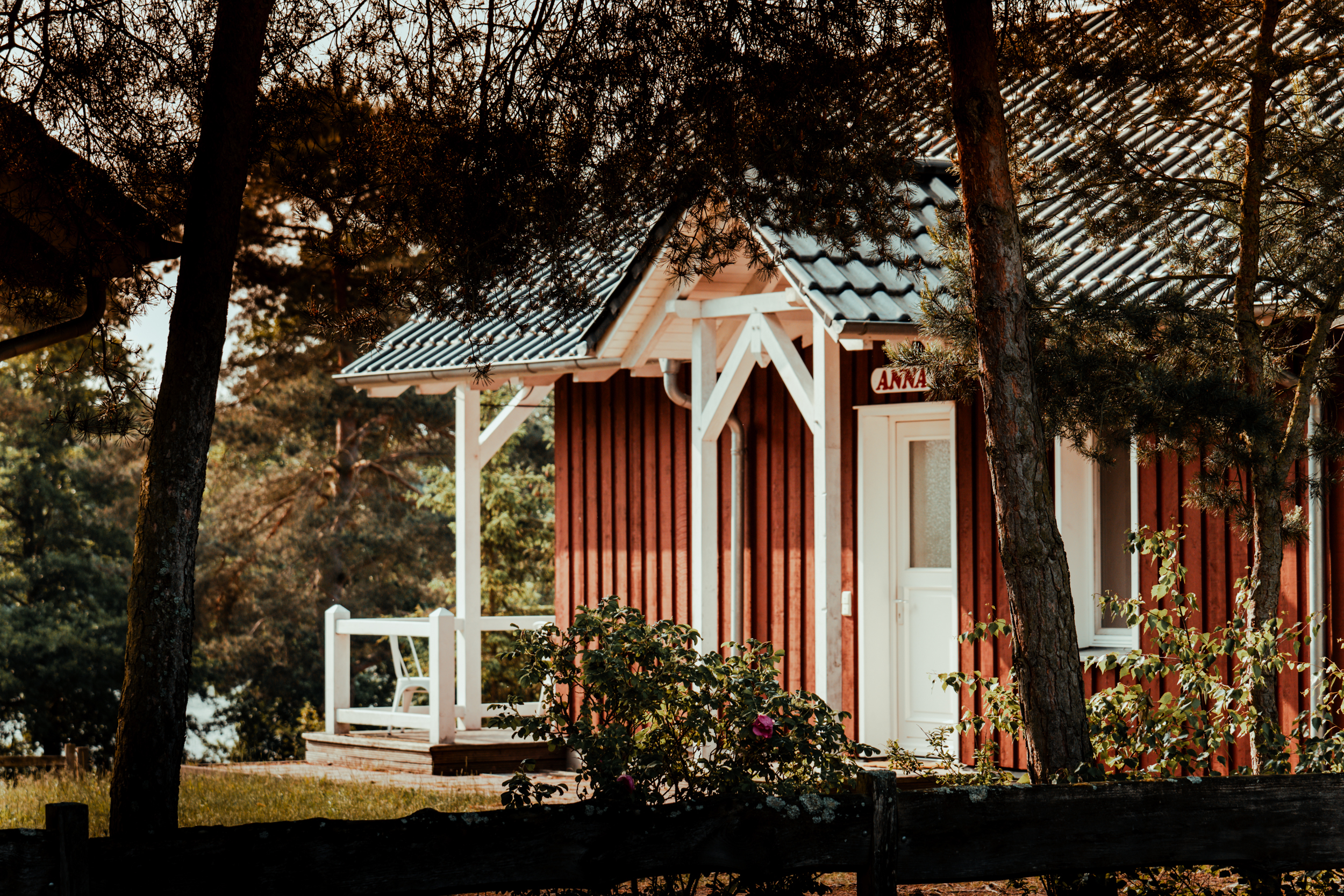 Eine rote Holzhütte mit weißer Tür und Veranda, umgeben von Bäumen und Grün, an einem sonnigen Tag – perfekt für „Mietunterkünfte Camping am See“.