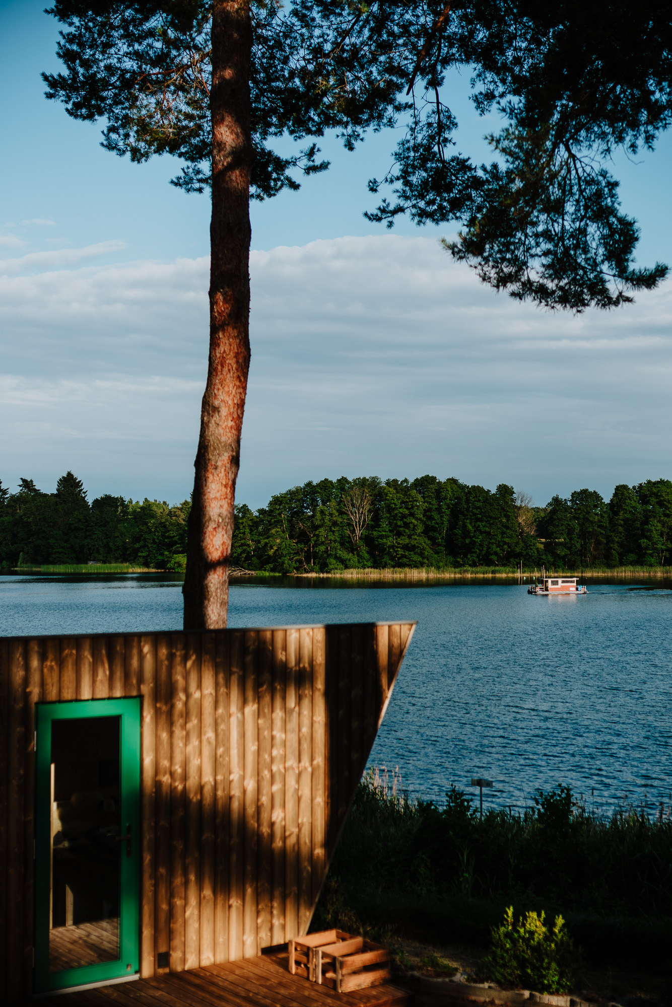 Eine Holzhütte am See, ein hoher Baum im Vordergrund und ein Boot auf dem Wasser mit Bäumen im Hintergrund.