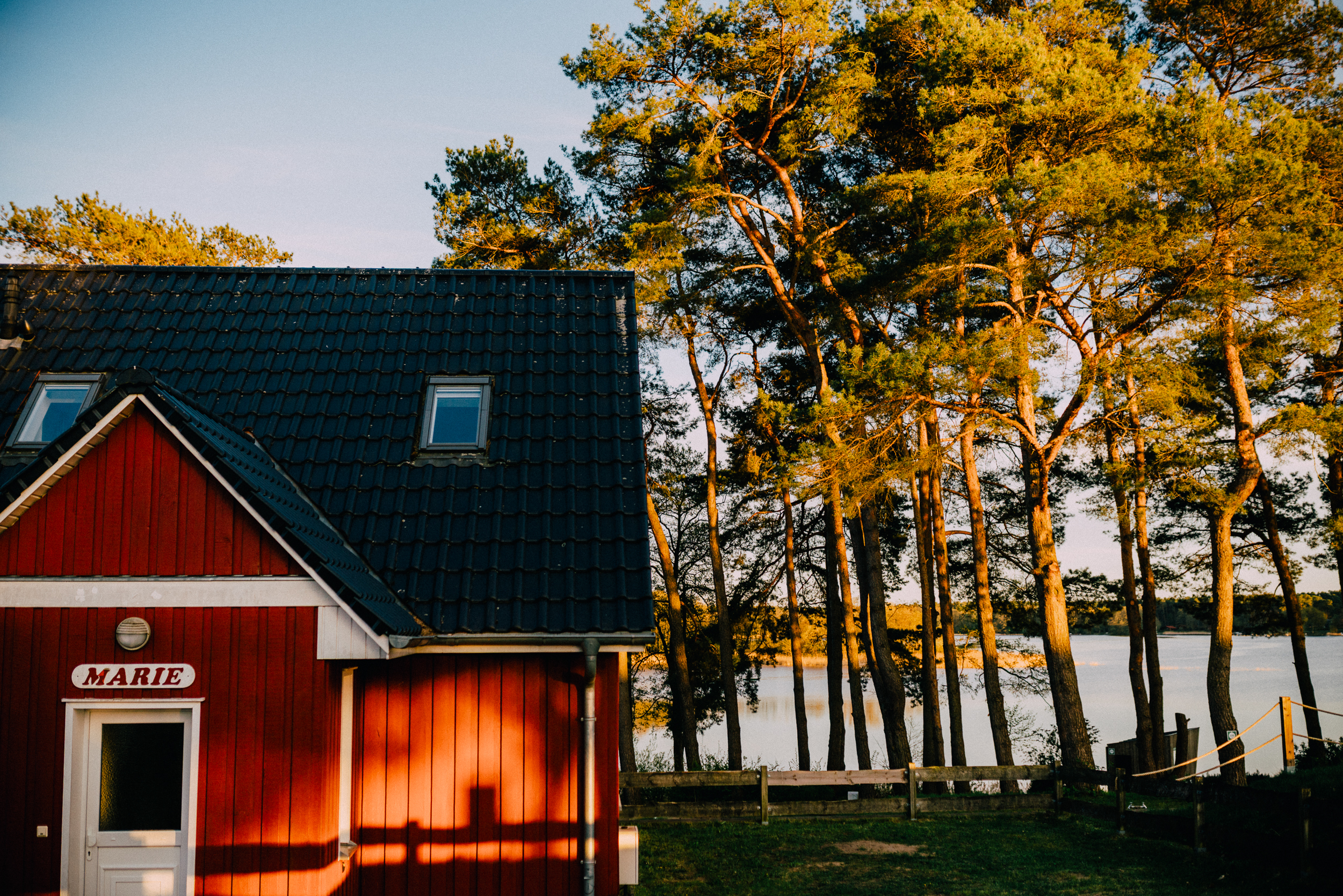 Ein rotes Haus mit der Aufschrift „Marie“ neben hohen Kiefern am Seeufer bei Sonnenuntergang, das den friedlichen Charme von „Mietunterkünfte Camping am See“ vermittelt.
